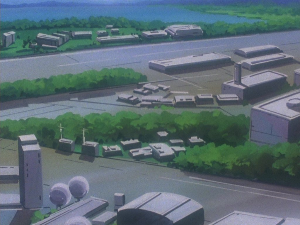Vista aérea de uma área urbana com edifícios modernos e vegetação ao redor, incluindo uma estrada e água ao fundo.