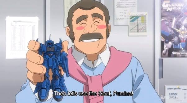 Homem sorridente segurando um modelo de robô azul chamado Gouf, em um ambiente de escritório, com papéis e cartazes ao fundo.